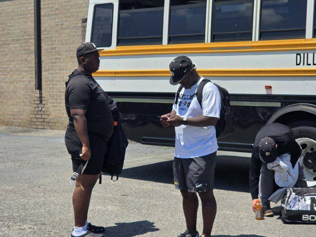 Three men stand near a school bus, two upright and one bending over a bag, wearing athletic clothes and hats on a sunny day.