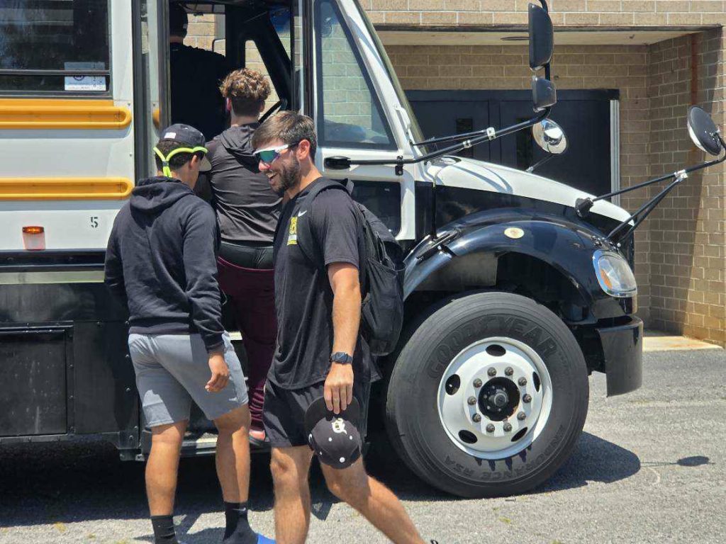 Three young men, dressed in athletic wear, board a white and black school bus parked near a brick building on a sunny day.