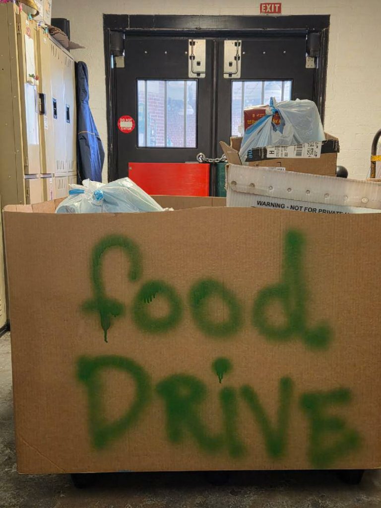 Large cardboard box labeled "food drive," filled with bags and boxes, sits in front of double doors in an indoor hallway.