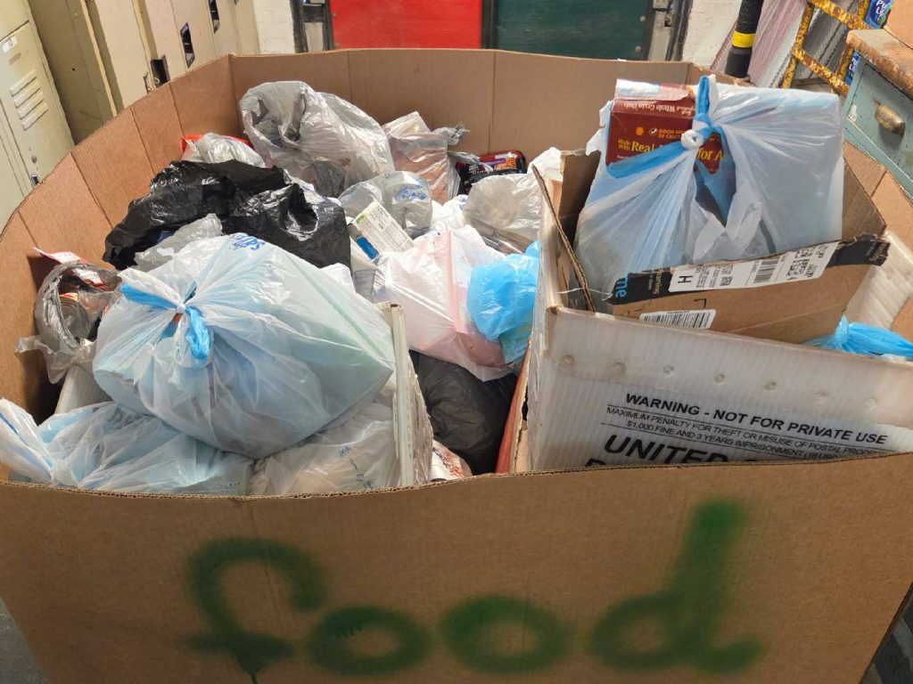 Large cardboard box labeled "food" filled with plastic bags and boxes containing donated food items, positioned in a storage or distribution area.