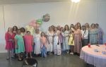 A group of women and girls pose together indoors, dressed in pastel dresses, with a balloon decoration and tea party setup in the background.