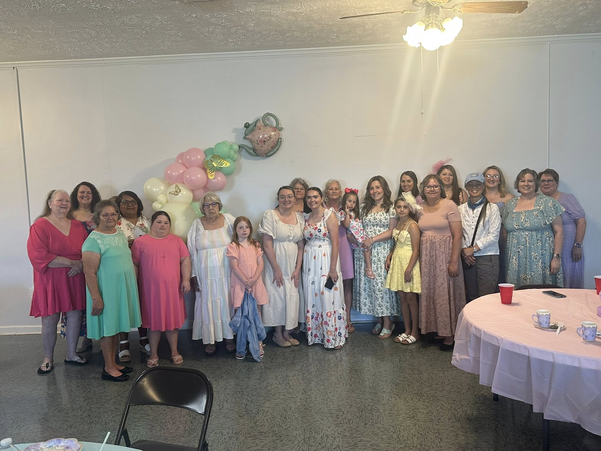 A group of women and girls pose together indoors, dressed in pastel dresses, with a balloon decoration and tea party setup in the background.