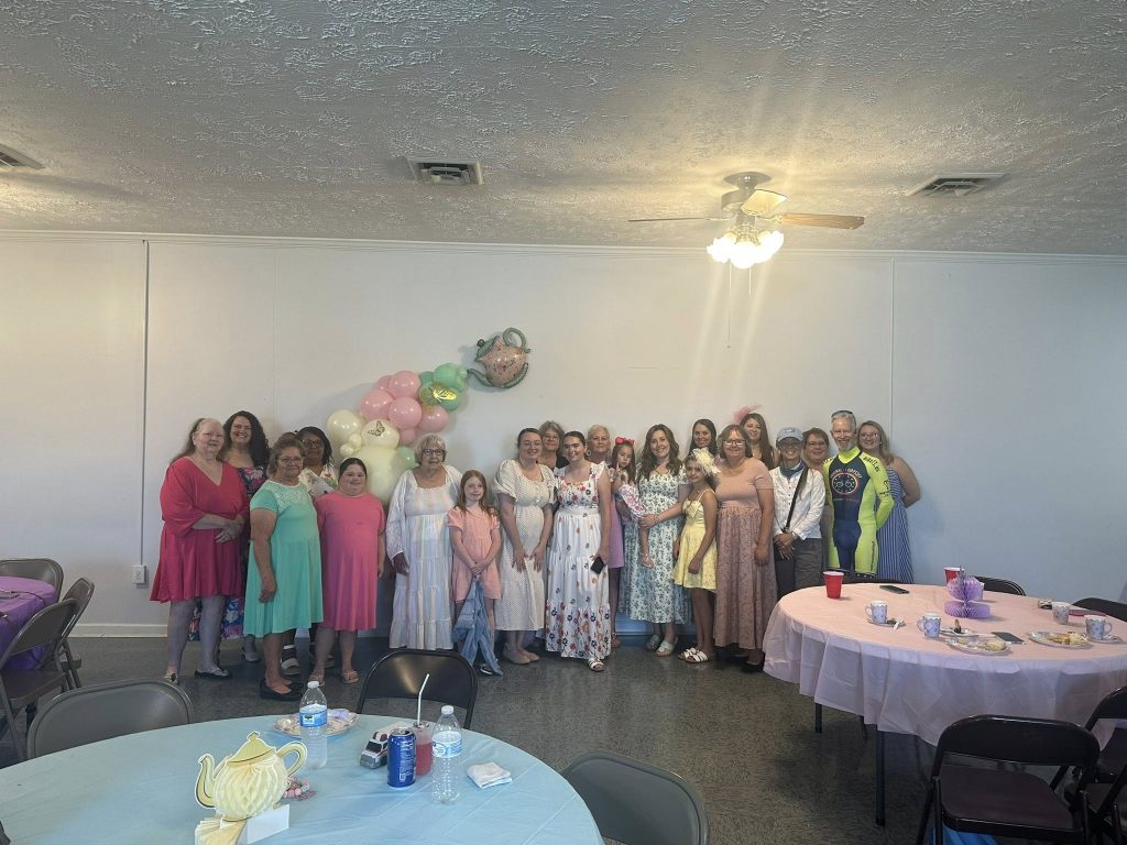 A group of people, mostly women and girls, pose in a decorated room with pastel balloons and tea-themed decor, dressed in colorful and semi-formal attire.