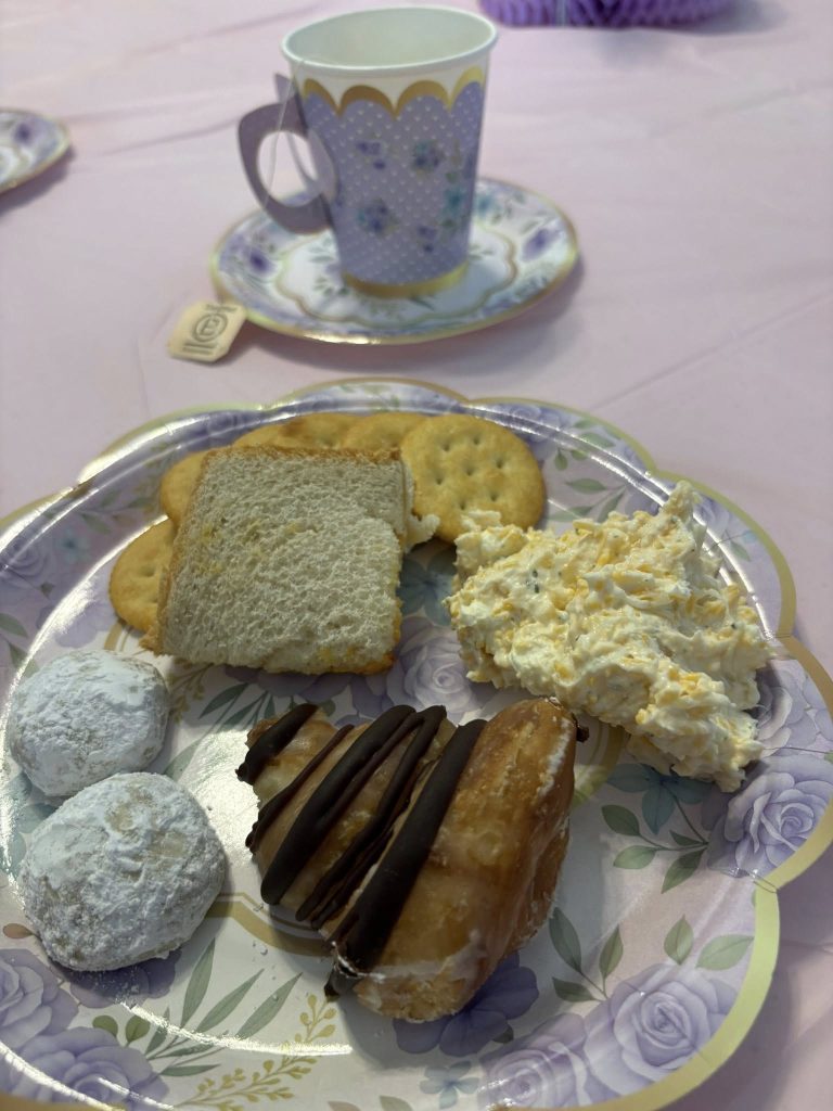 Paper plate with cookies, crackers, bread, cheese spread, and a chocolate pastry. Floral teacup and saucer with tea bag on a pastel tablecloth in the background.