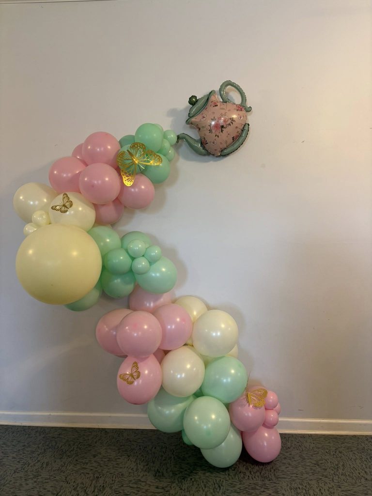 Pastel balloon arrangement flowing from a floral teapot decoration, adorned with gold butterfly accents, against a plain wall.