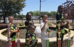 Five children in uniforms pose proudly with large trophies in front of a fountain on a sunny day.