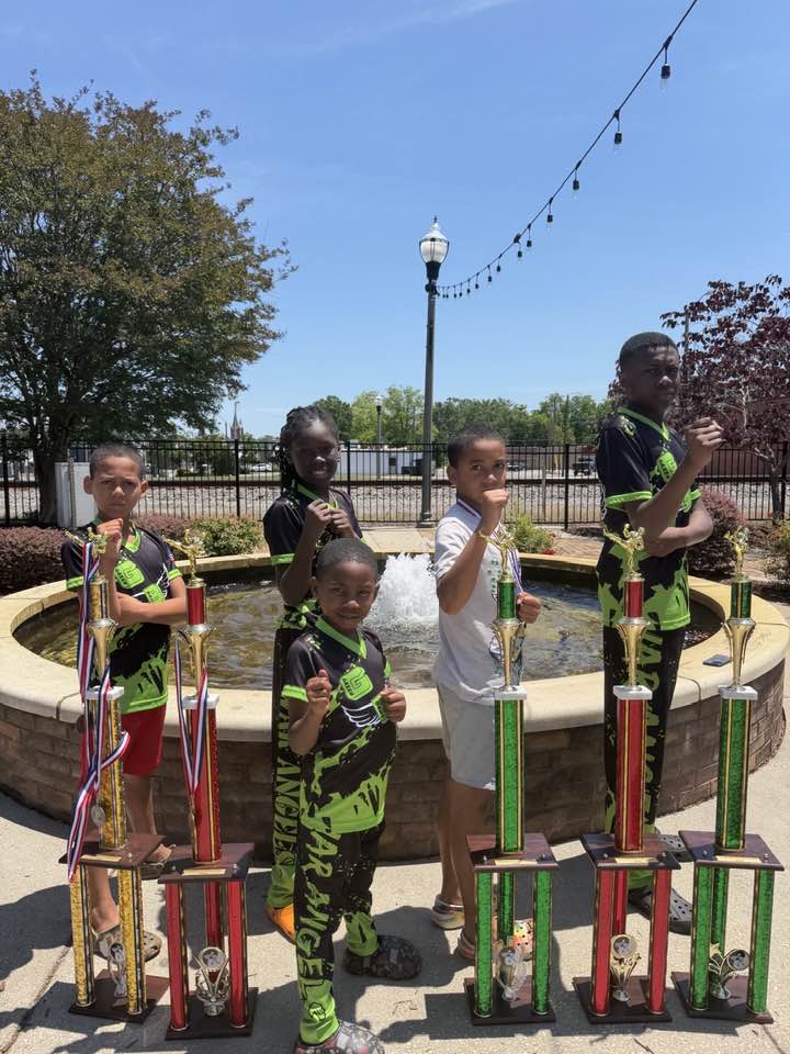 Five children in uniforms pose proudly with large trophies in front of a fountain on a sunny day.