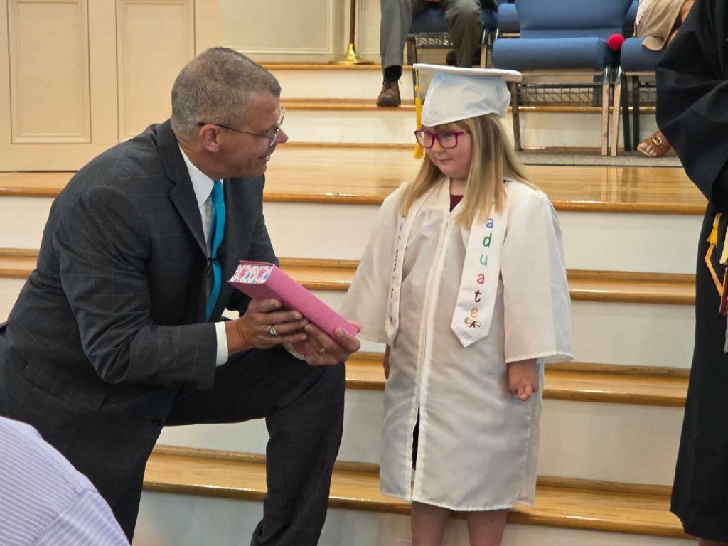 A man in a suit kneels, smiling and handing a diploma to a young girl in a graduation cap and gown on wooden steps.