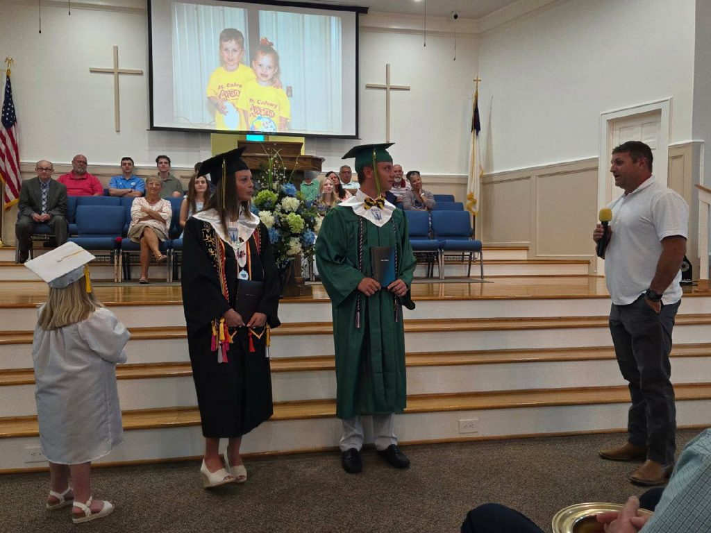 Three graduates stand at the front of a church as an adult speaks to them, with people seated behind and a childhood photo projected above.