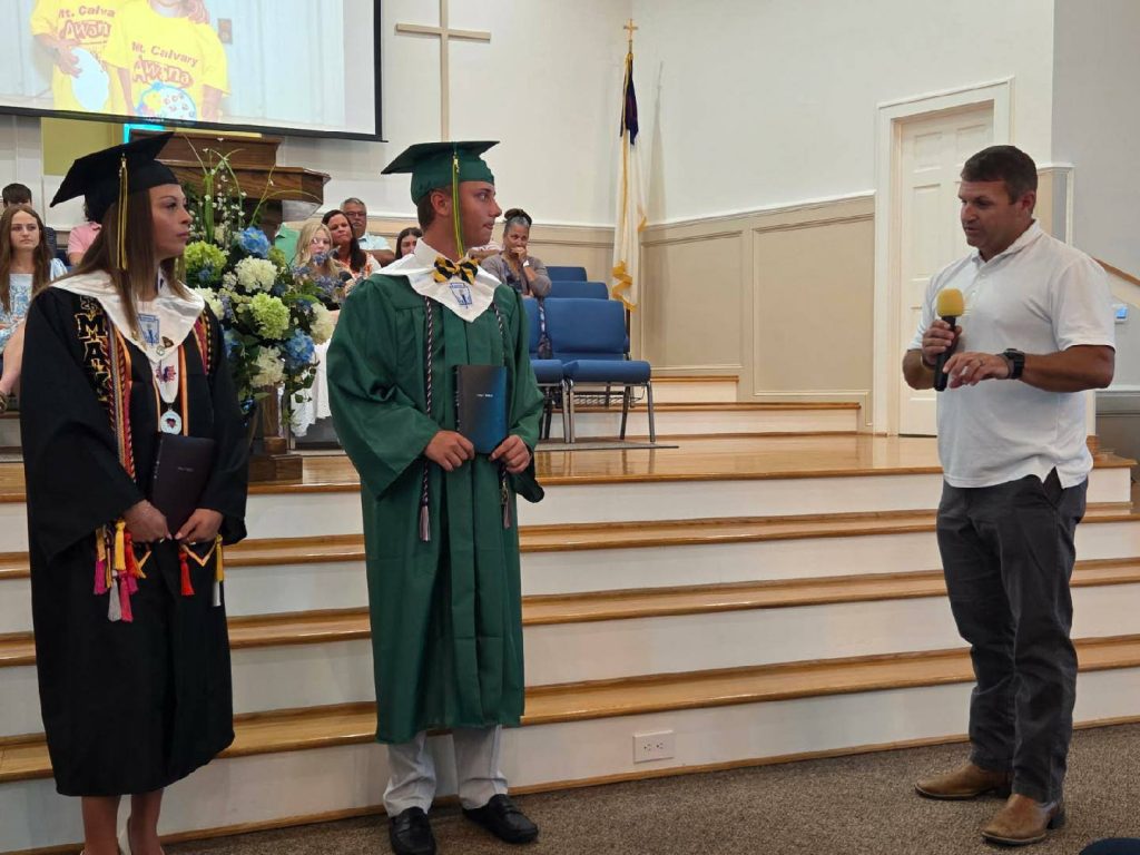 Two graduates in caps and gowns stand on stage while a man speaks into a microphone in a church setting.
