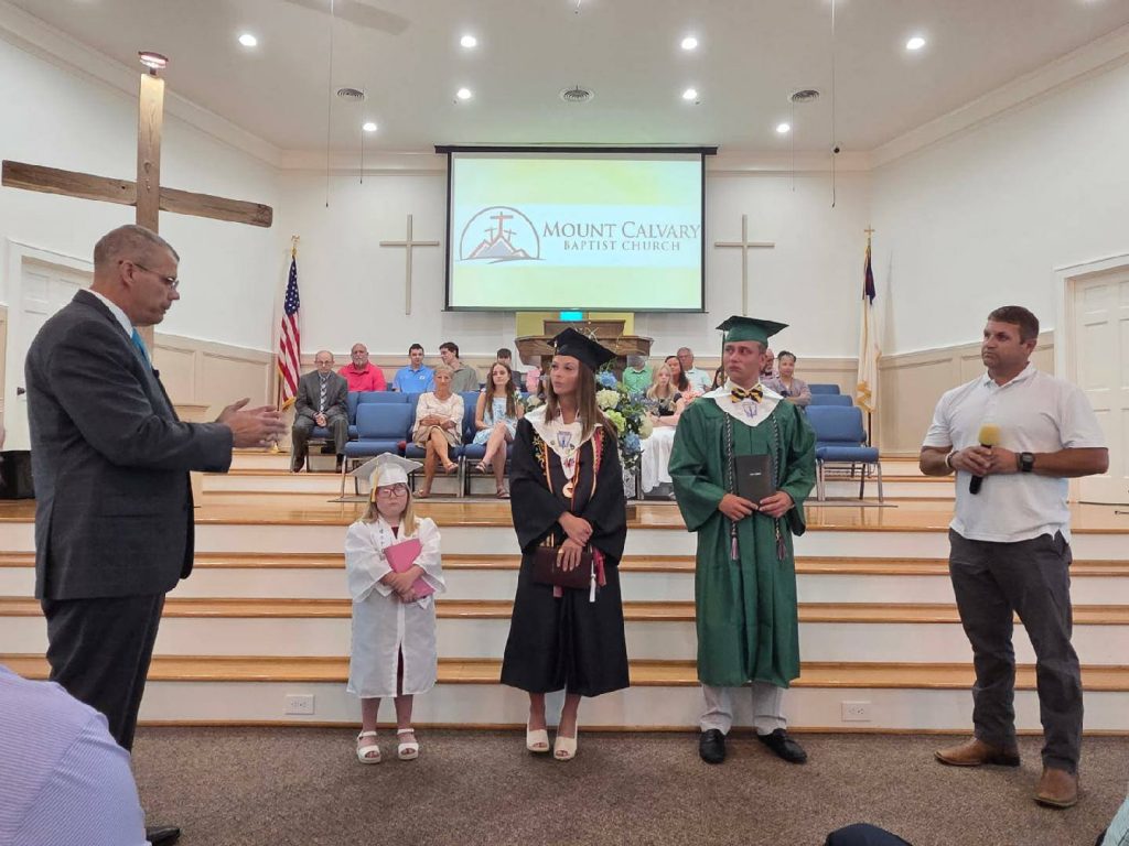 Three graduates in caps and gowns stand in a church with two adults, as people watch from the background.