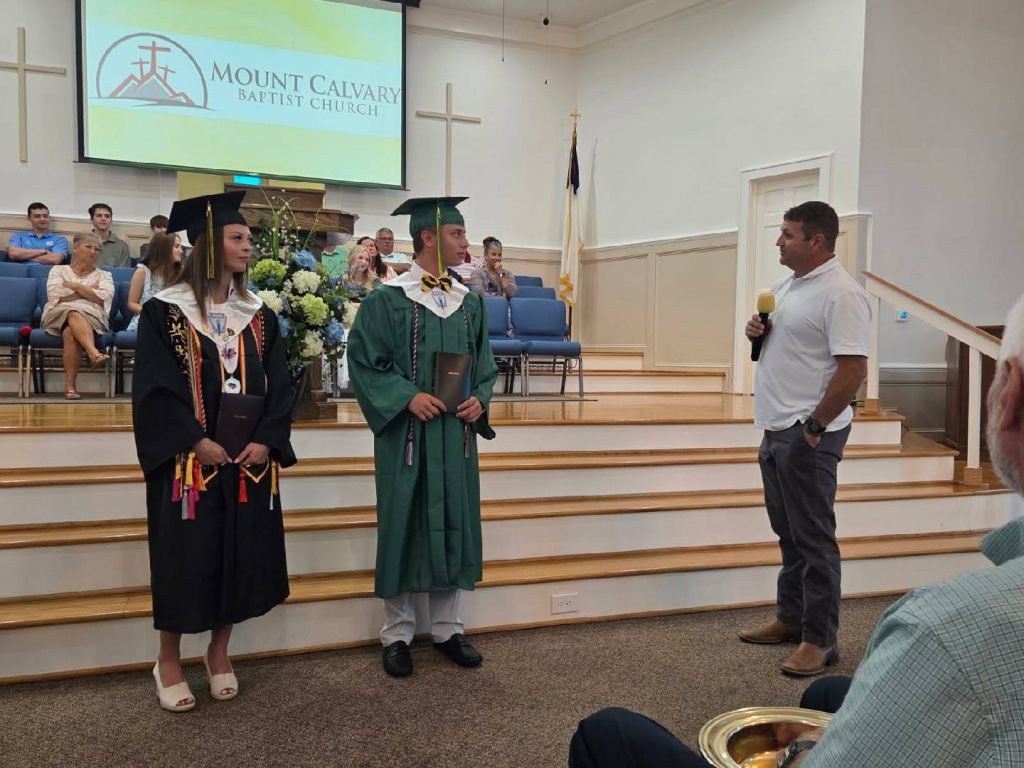 Two graduates in caps and gowns stand at the front of a church while a man speaks to them with a microphone.