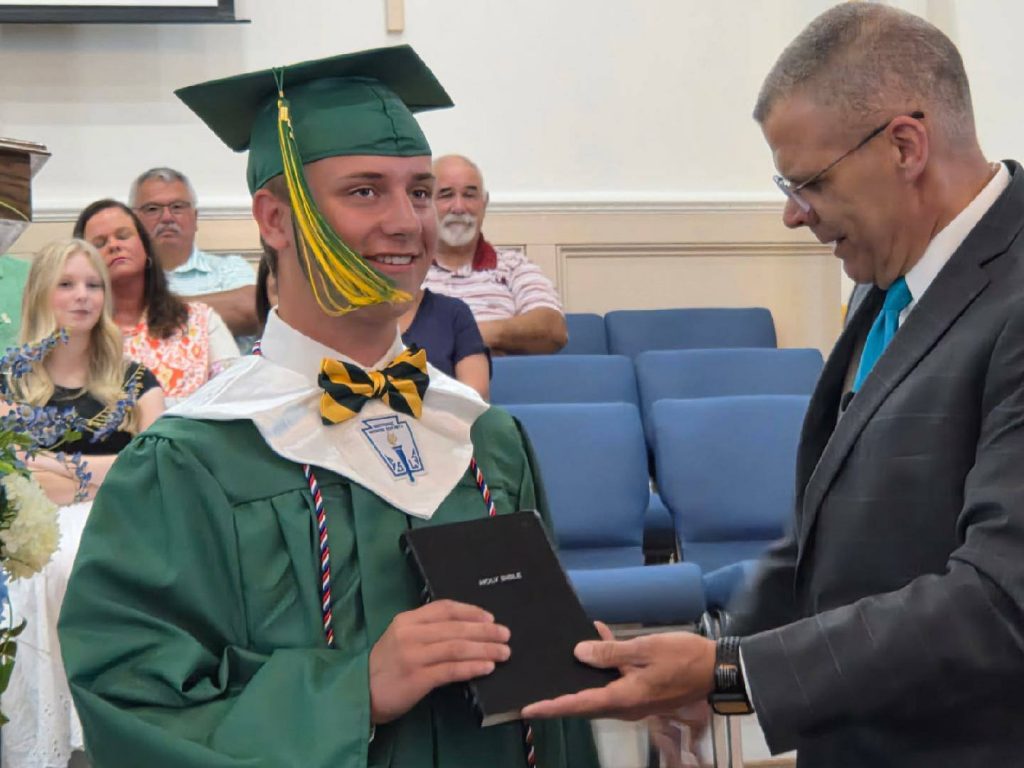 A smiling graduate in a green cap and gown receives a Bible from an older man in a suit, with people seated in the background.
