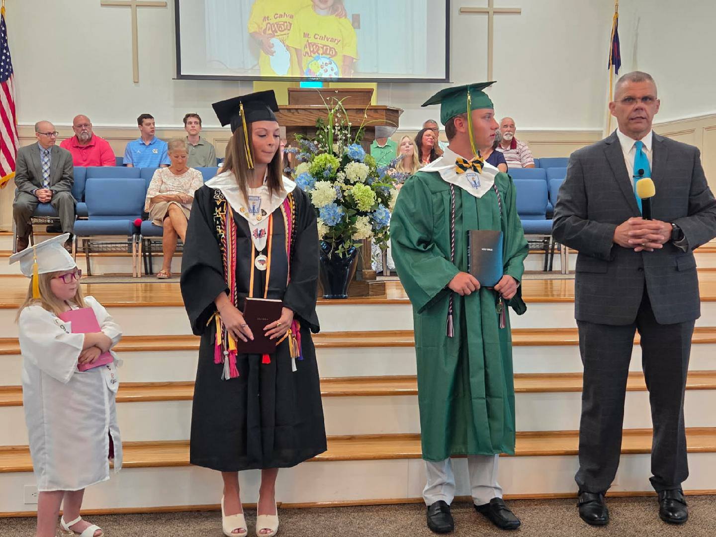 Three graduates and a man in a suit stand at the front of a church, with people seated behind them and a large bouquet of flowers nearby.