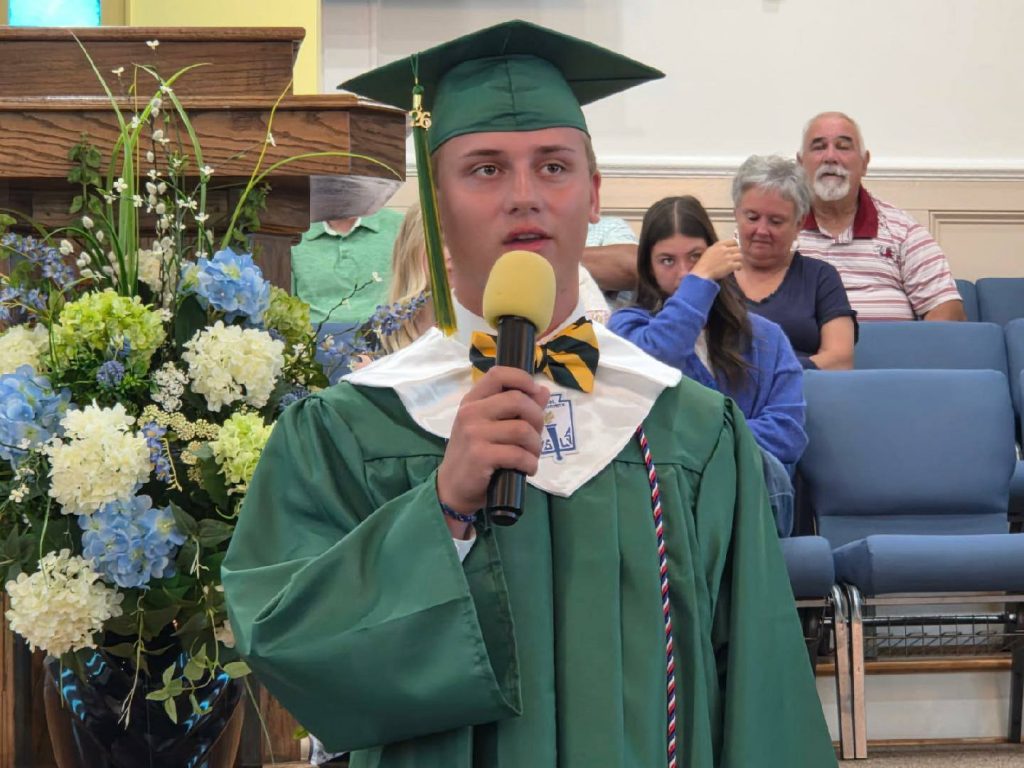 A graduate in a green cap and gown speaks into a microphone at a ceremony, with flowers and an audience in the background.