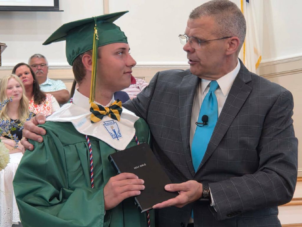 A graduate in a green cap and gown receives a Holy Bible from a man in a suit during a ceremony, with people seated in the background.
