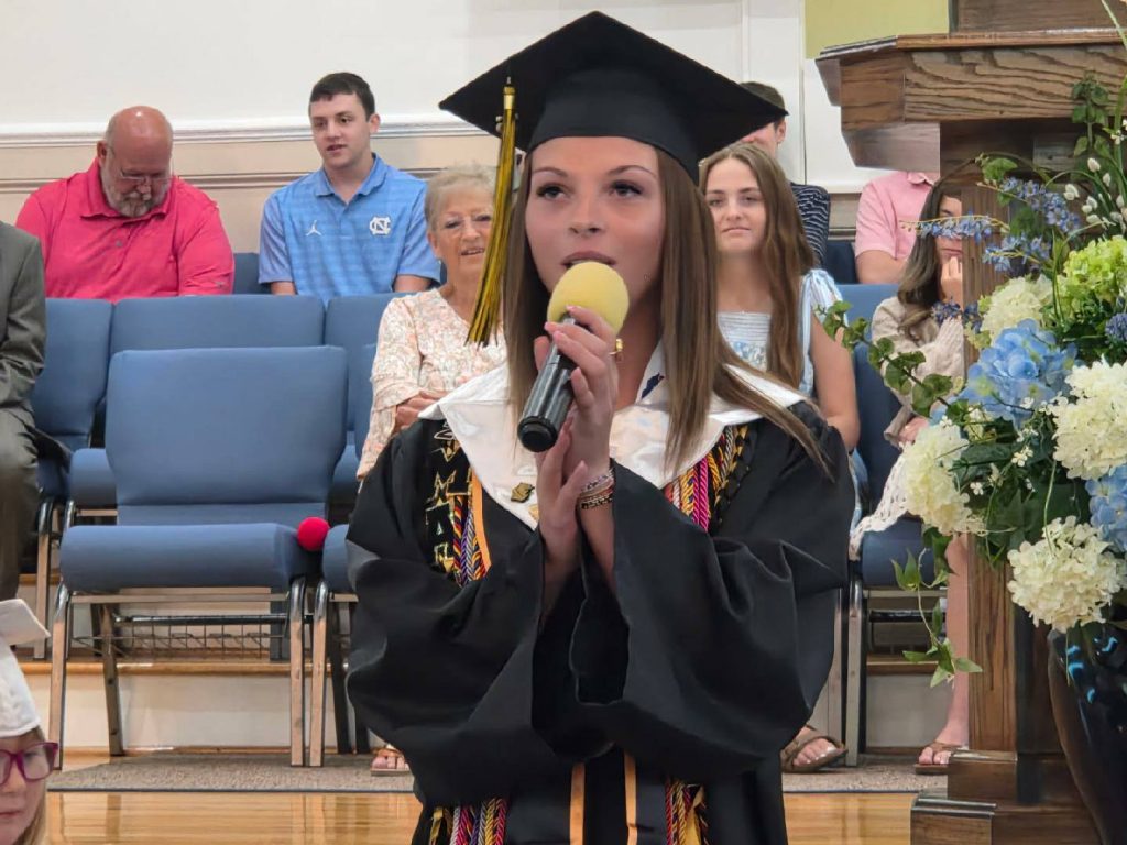 Young woman in graduation cap and gown speaks into a microphone during a ceremony, with people seated in the background and a large floral arrangement nearby.