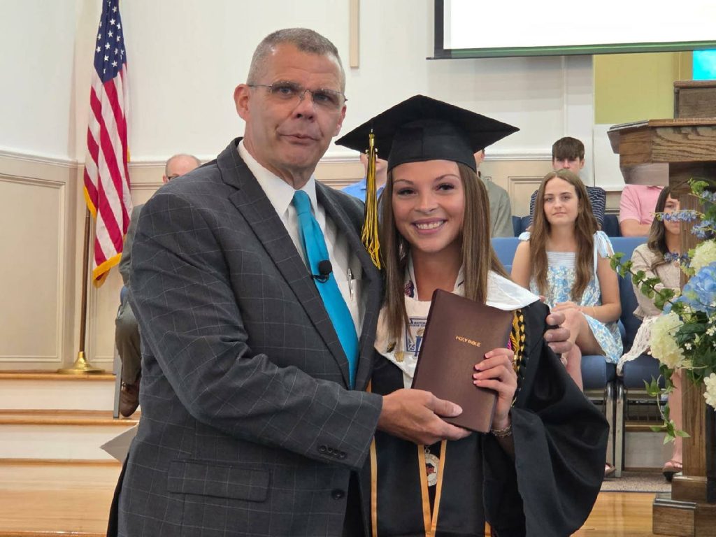 A man in a suit stands beside a smiling female graduate holding a Bible, with people seated and an American flag in the background.
