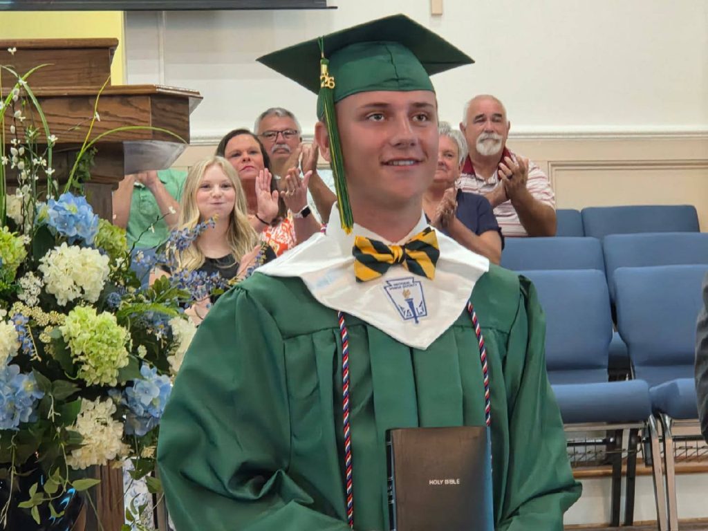 Young graduate in green cap and gown holding a Bible, standing in front of seated audience clapping, with floral arrangement nearby.