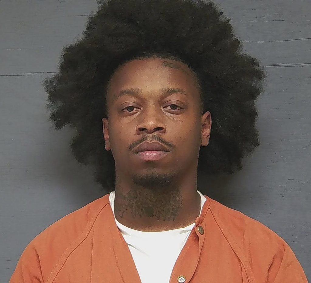 A man with an afro hairstyle wearing an orange prison jumpsuit poses for a mugshot against a plain gray background.