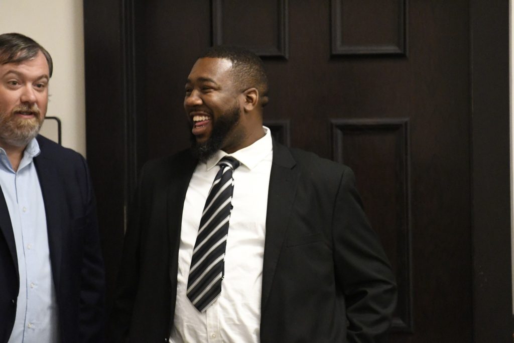 Two men in suits stand indoors, one smiling and laughing, the other looking ahead, in front of a dark wooden door.