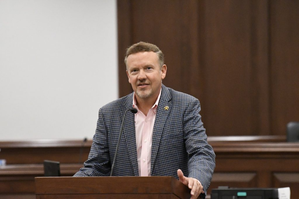 A man in a checked blazer speaks at a podium in a formal, wood-paneled room with a microphone in front of him.