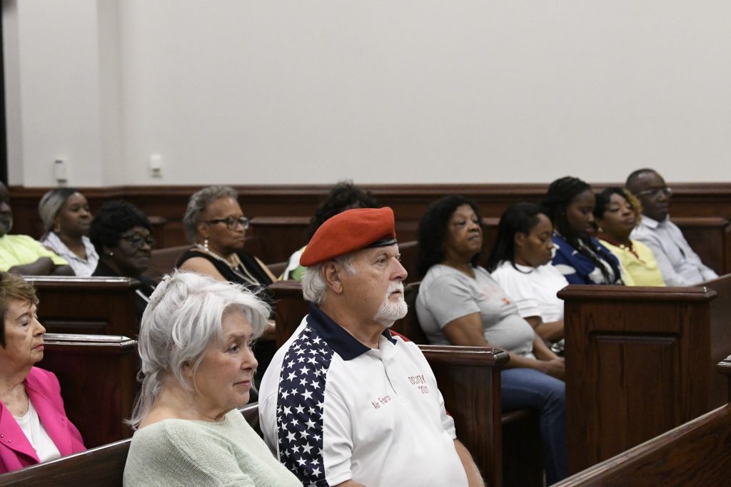 People seated in wooden benches attentively listening in a courtroom or public meeting, including a man wearing a red beret and a star-spangled shirt.