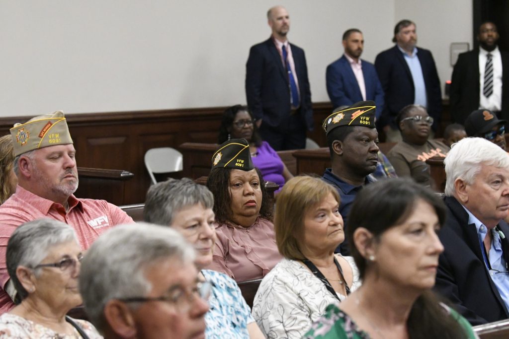 People, including veterans in VFW caps, sit attentively in a courtroom or meeting hall, with others standing in the background, attending an event or ceremony.