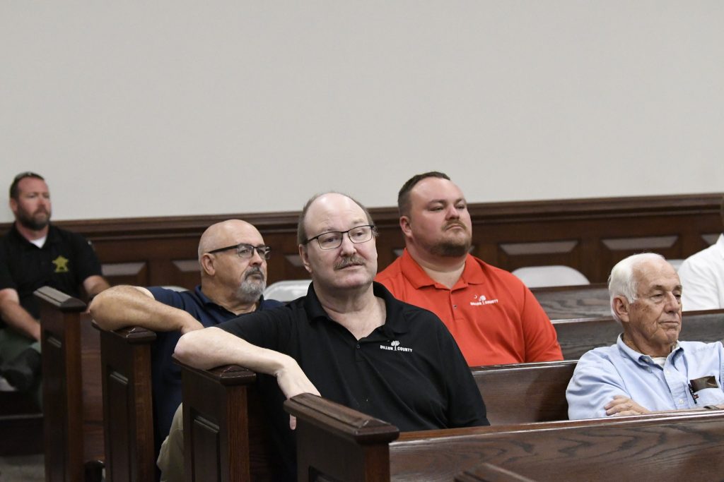Several men sit in wooden benches, appearing attentive in a courtroom or meeting room with neutral walls and sparse decor.