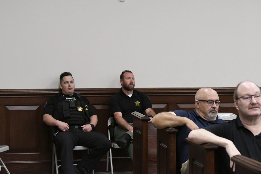 Two sheriff officers and two men sit on chairs and benches in a courtroom, facing forward with neutral expressions.