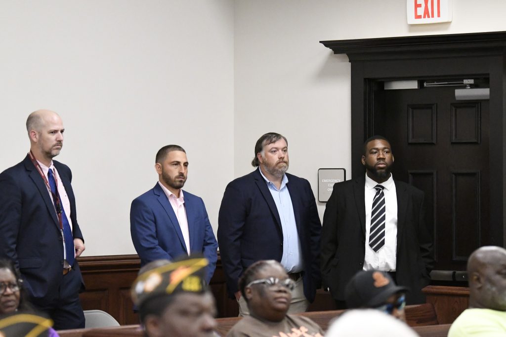 Four men in suits stand in a courtroom near an exit door, with seated audience members visible in the foreground.