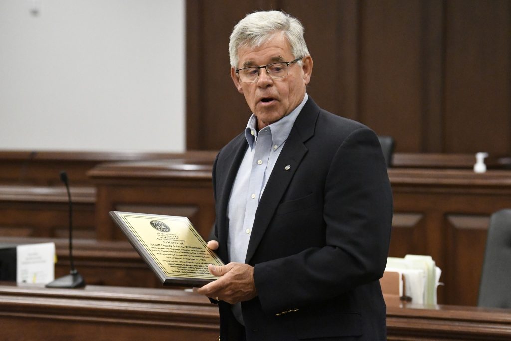 older man in a suit stands in a courtroom holding a plaque, appearing to speak during an award or recognition event.