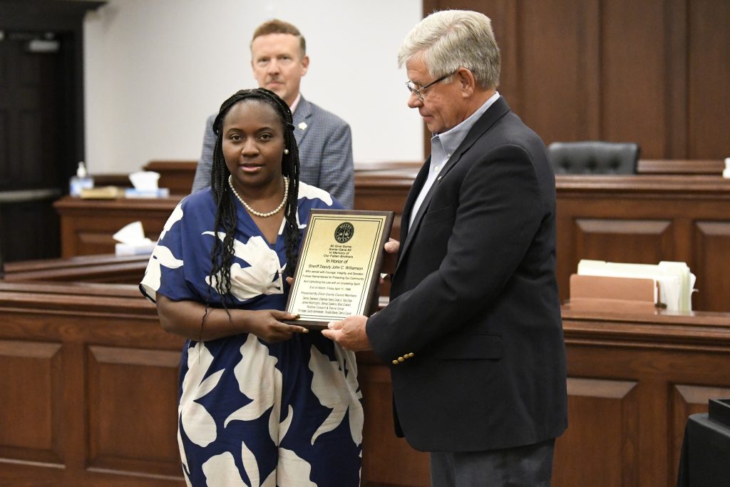 A woman and man stand in a courtroom holding a plaque together, while another man stands in the background watching the presentation.