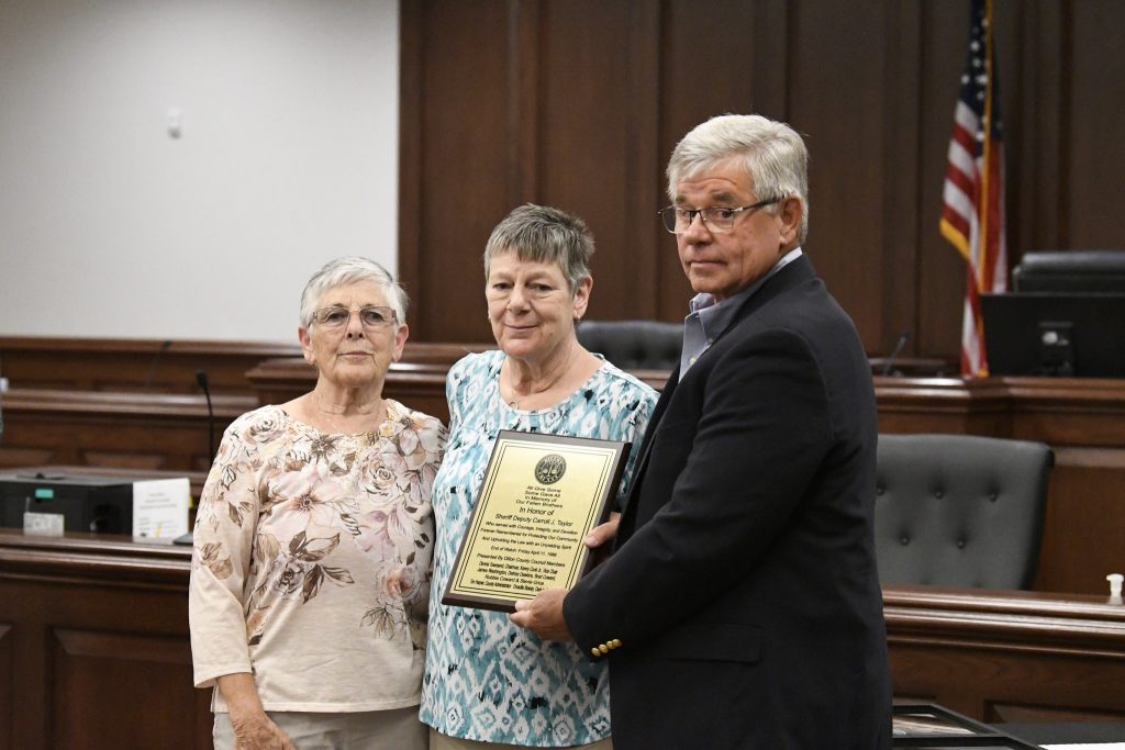 Three people standing in a courtroom, one holding a plaque, with an American flag visible in the background.