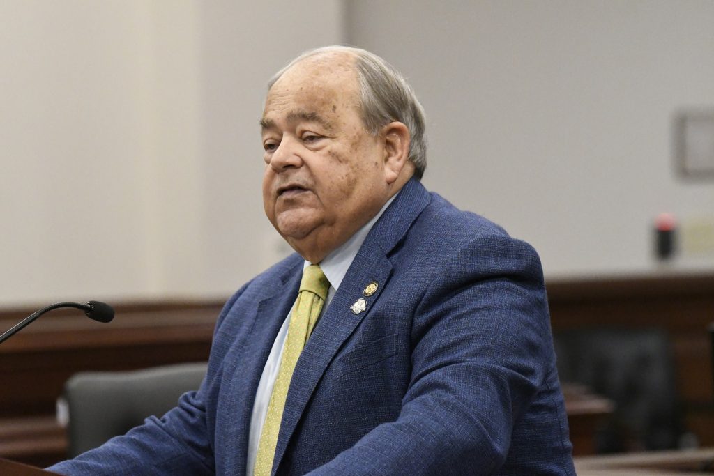 Older man in a blue suit and yellow tie speaks at a podium in a formal indoor setting, possibly a courtroom or government building.