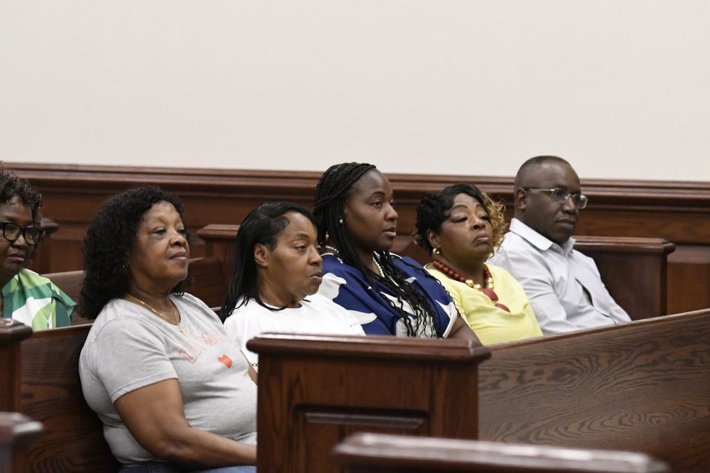 A group of adults sit quietly in a courtroom pew, listening attentively during a legal proceeding.