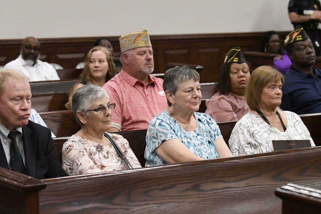 People sit in wooden pews inside a courtroom, some wearing veterans’ hats, with serious and attentive expressions during a formal event or proceeding.