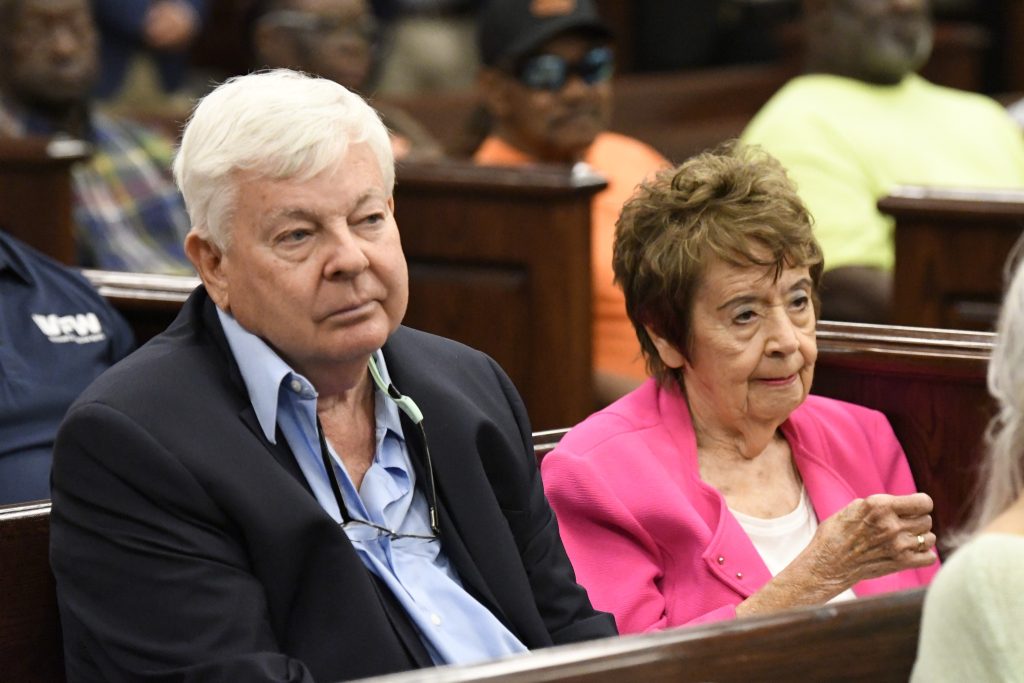 elderly man and woman sit together in a crowded room, attentively listening. The woman wears a pink jacket, and the man wears a suit jacket and light blue shirt.