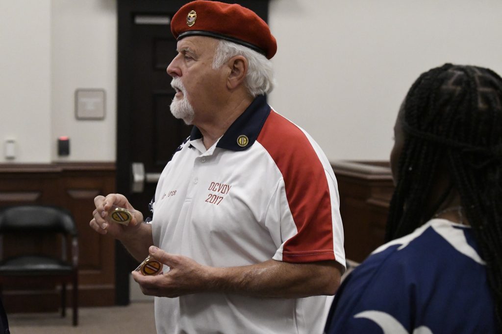 Older man in a red beret and white shirt holding medallions, speaking to a woman in a room with wooden furnishings.