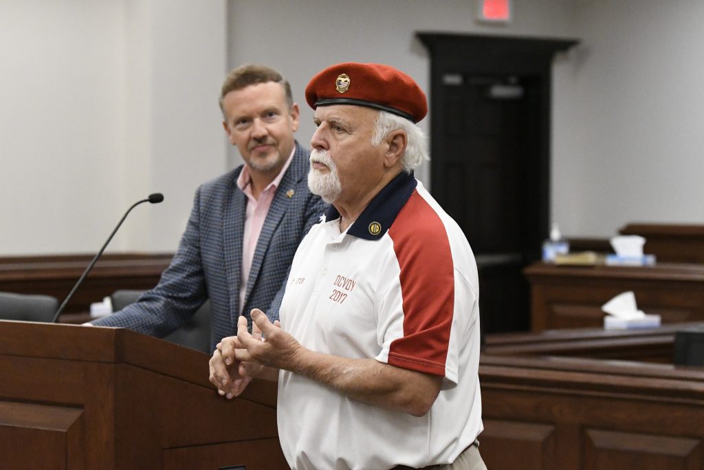 Two men standing in a courtroom, one wearing a red beret and casual polo, the other in a checked suit, speaking at a wooden podium.