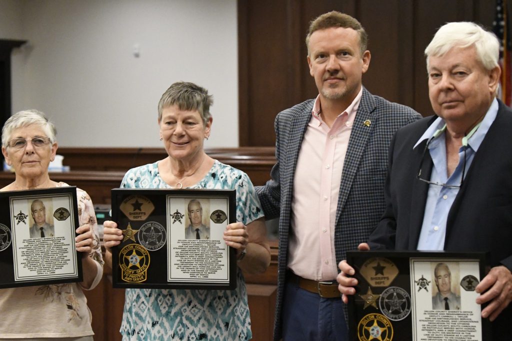 Four adults stand together, three holding framed plaques with a man’s photo and sheriff’s department insignia, in a formal indoor setting.