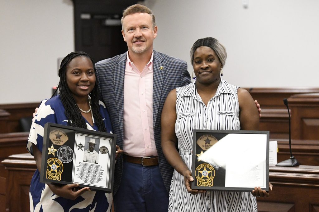 Three people stand together in a courtroom, two women holding framed plaques with sheriff department badges and text, smiling for a recognition or award ceremony.