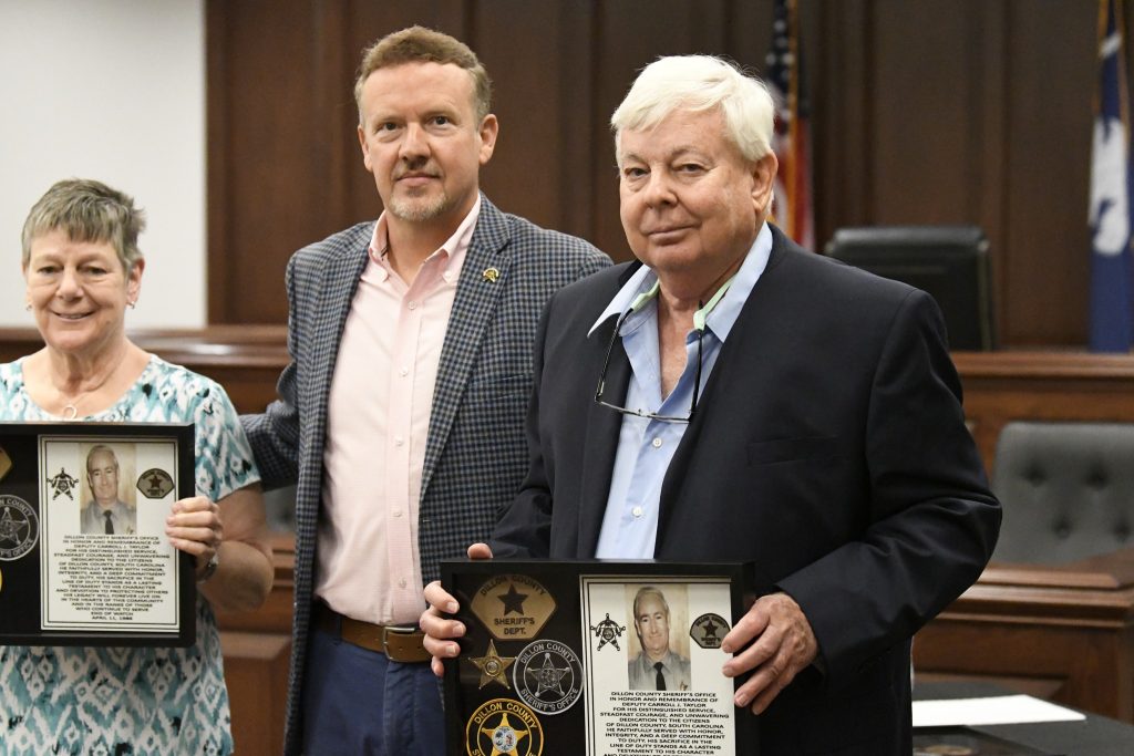 Three people standing in a courtroom, two holding framed plaques with photographs and badges, appearing to receive awards or recognition.
