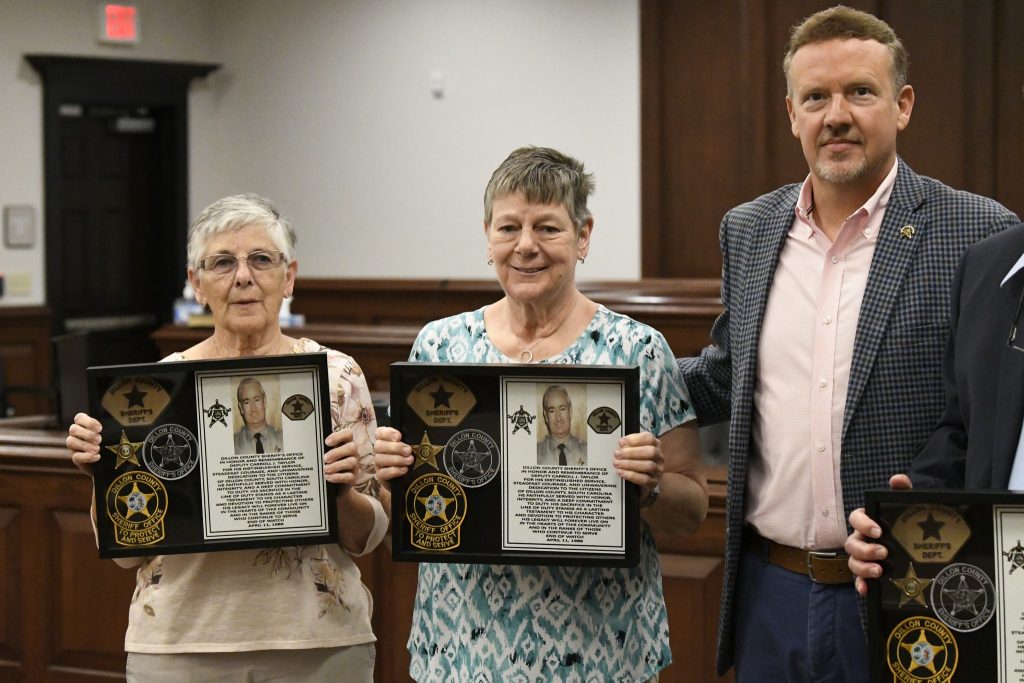 Two women hold framed sheriff’s department awards in a ceremony, standing next to a man in a checked blazer inside a wood-paneled room.