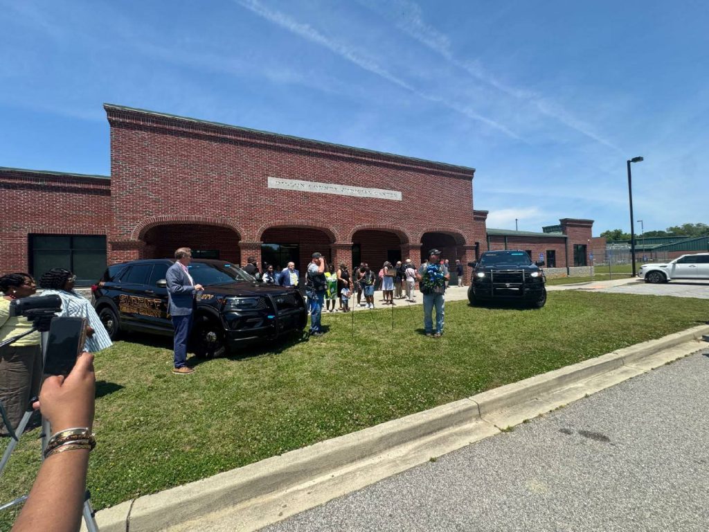 People gathered outside a brick sheriff’s office building, with two black sheriff vehicles parked on the grass under a clear blue sky.
