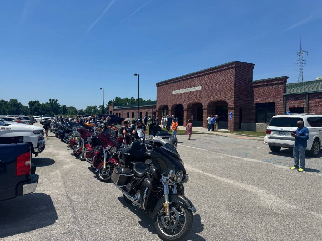 A row of parked motorcycles and people outside a brick building under a clear blue sky.