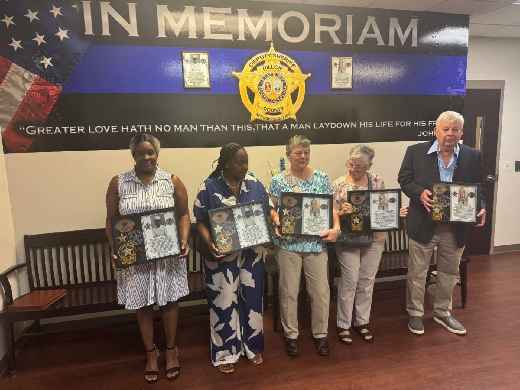 Five people stand holding framed memorial plaques in front of a wall that says "IN MEMORIAM" at a sheriff's office, honoring fallen officers.