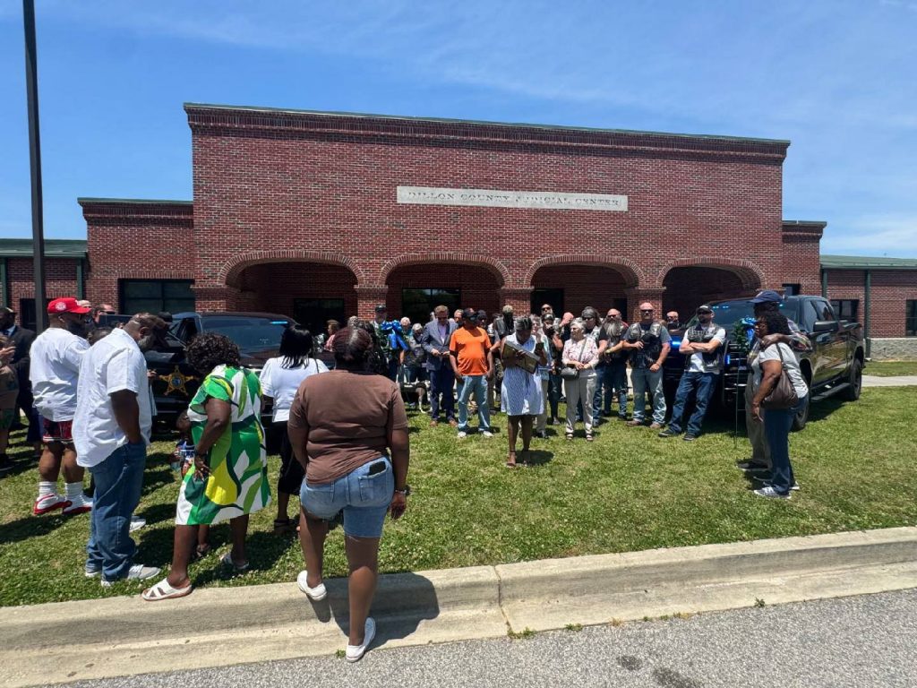 A group of people gathers outside a brick building labeled "Dillon County Judicial Center" on a sunny day, some standing near parked vehicles.