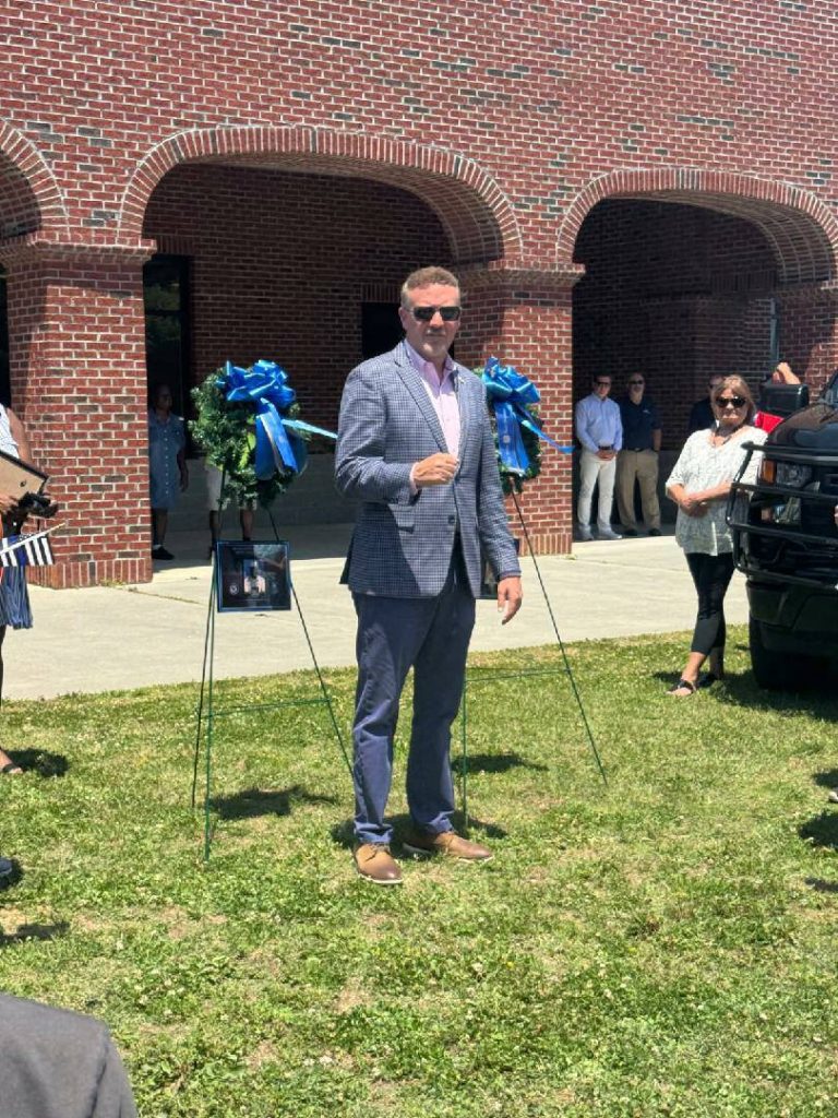 A man in sunglasses and a suit stands on grass near a brick building, with people, wreaths, and a photo on display around him.