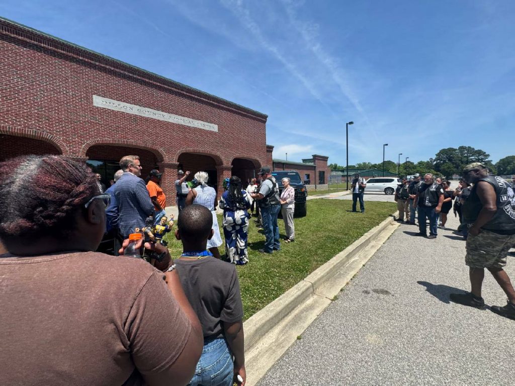A group of people gather outside a red brick building under a sunny sky, some taking photos and wearing casual clothing.